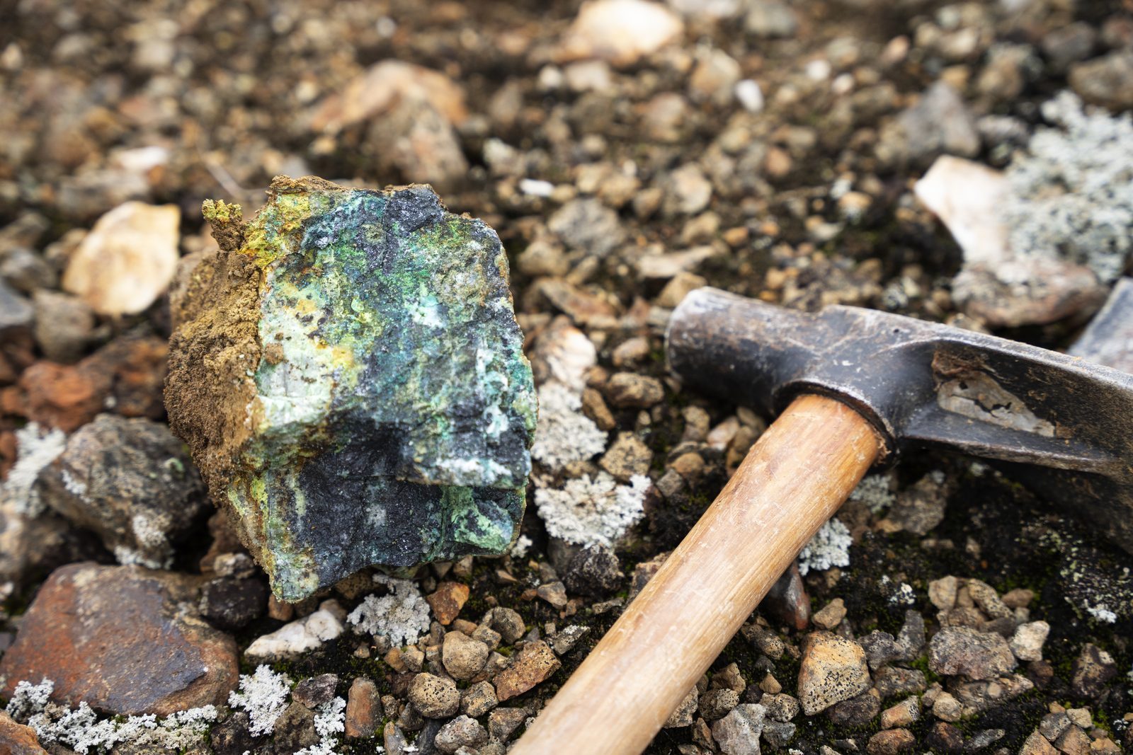 A copper-mineralized rock specimen, green with malachite, resting on scree beside a geologist's rock hammer