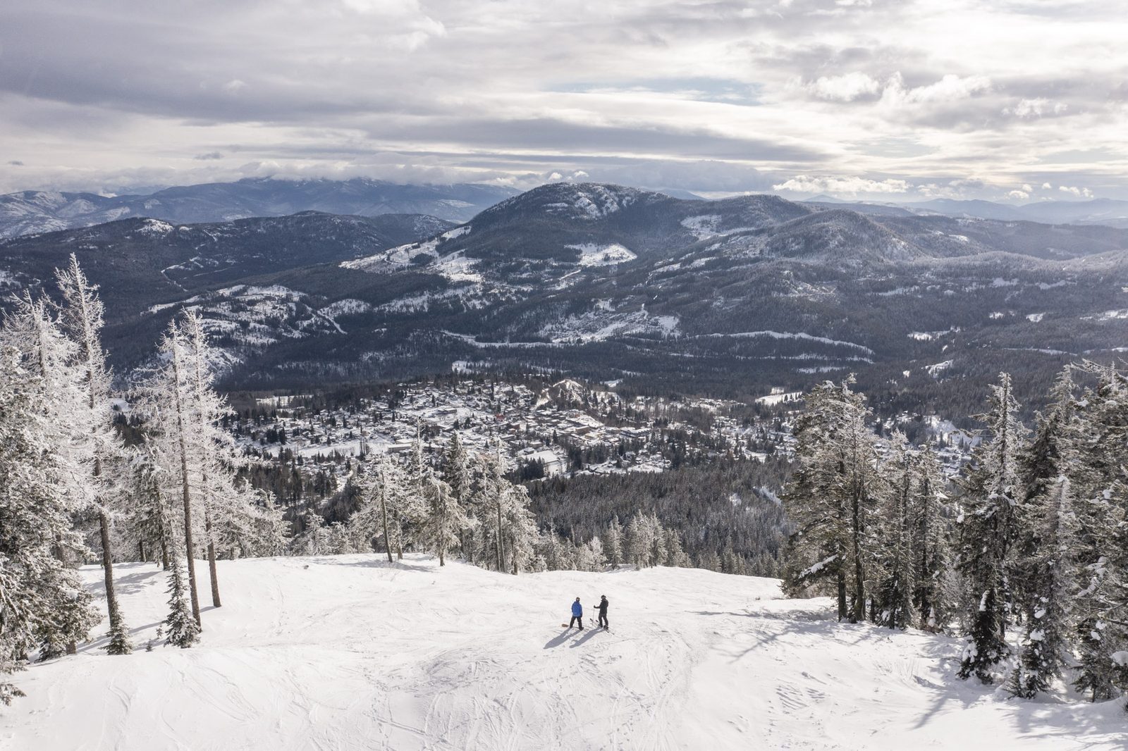 Two skiers pause on a run high on Red Mountain, with the town of Rossland visible far below in the valley